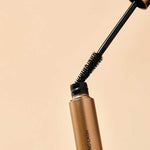 The INIKA Organic Long Lash Mascara 8ml, in a gold tube with a black wand and bristled brush partly removed from the container, is set against a beige background.