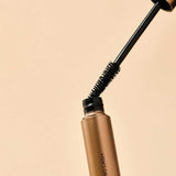 The INIKA Organic Long Lash Mascara 8ml, in a gold tube with a black wand and bristled brush partly removed from the container, is set against a beige background.