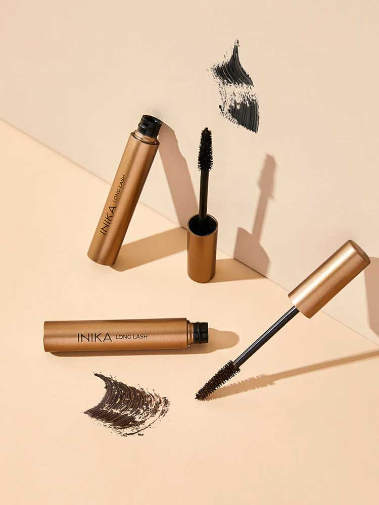 Three gold tubes of INIKA Organic Long Lash Mascara 8ml are displayed on a beige surface, two with their wands open and visible. Black mascara brush strokes are placed beside each tube.