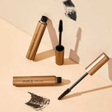 Three gold tubes of INIKA Organic Long Lash Mascara 8ml are displayed on a beige surface, two with their wands open and visible. Black mascara brush strokes are placed beside each tube.
