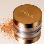 An INIKA Organic Loose Mineral Bronzer - Sunkissed 8g with a round gold lid rests on a light surface, its bronzing powder spilled beside the container.