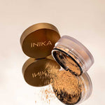 An open INIKA Organic Loose Mineral Bronzer - Sunkissed 8g with a gold lid rests on a reflective surface, some bronzer spilling out. The propped lid beside the round container clearly displays the INIKA brand name.
