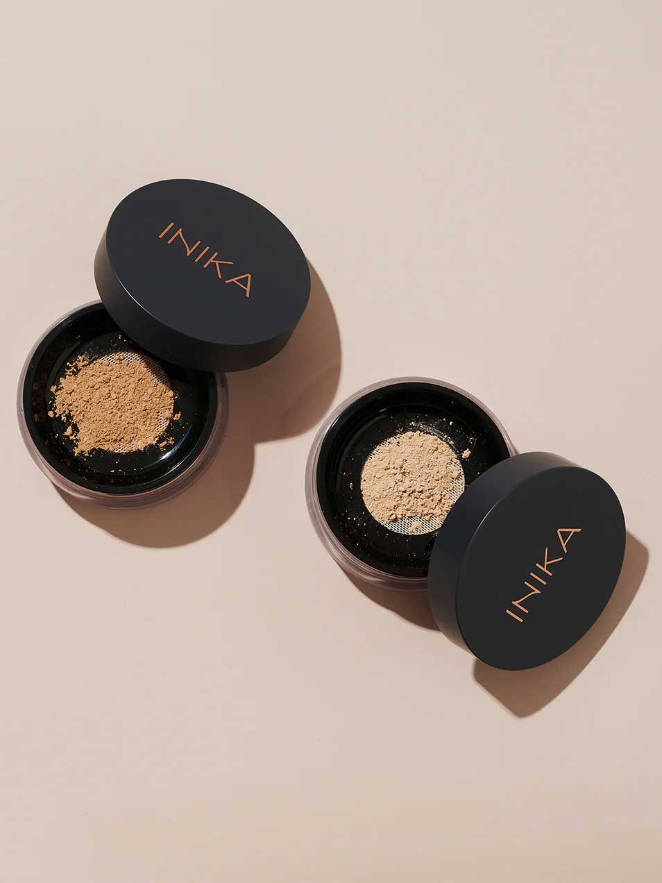 Two round containers of INIKA Organic Loose Mineral Foundation SPF25 8g with black lids sit on a beige surface. One contains a darker shade, the other a lighter one, both offering customizable organic coverage.