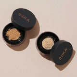 Two round containers of INIKA Organic Loose Mineral Foundation SPF25 8g with black lids sit on a beige surface. One contains a darker shade, the other a lighter one, both offering customizable organic coverage.