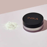 A small pile of loose white INIKA Organic Mattify Mineral Setting Powder 7g sits next to its round container with a black lid labeled "INIKA" in copper, placed on a light pink and white surface.