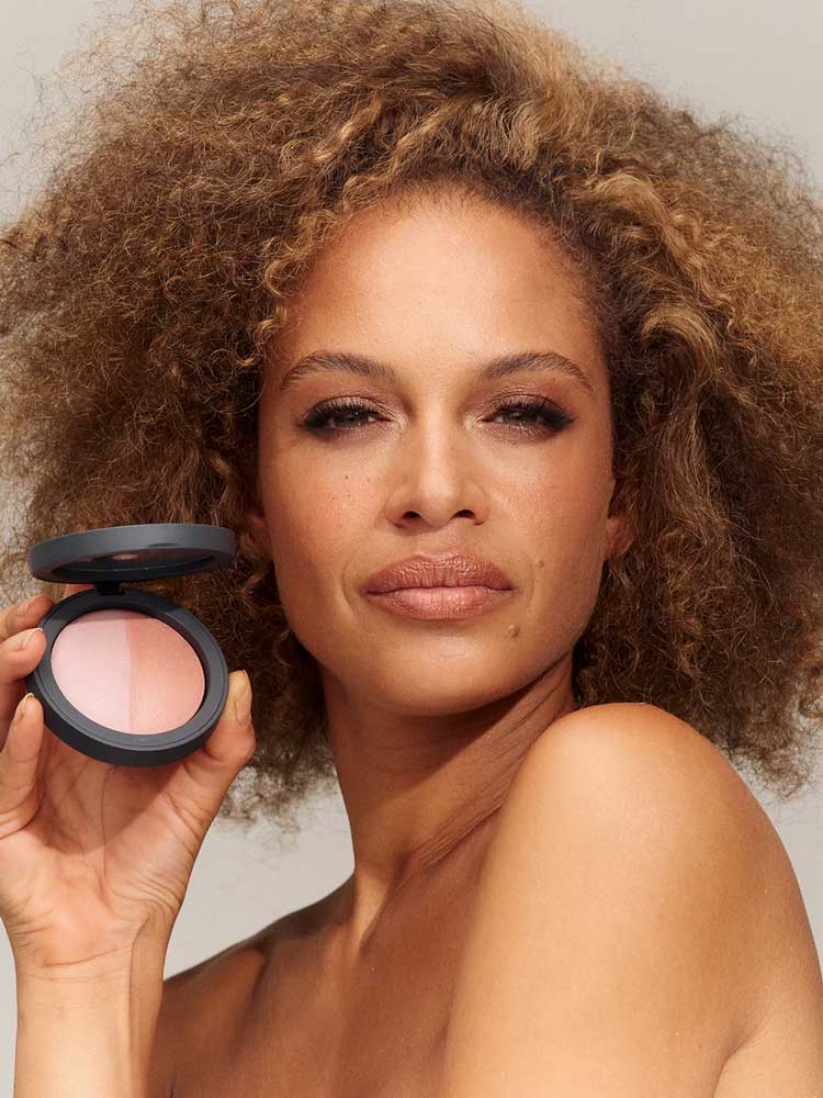 A woman with curly hair holds the INIKA Organic Mineral Baked Blush Duo - Pink Tickle 6.5g near her face, showing off the vegan blush. She has a neutral look and natural makeup against a plain, light background.