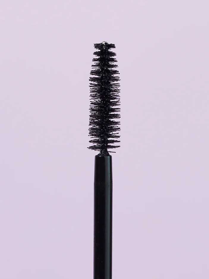 A close-up of the INIKA Organic Purity Lash Mascara - Black 8.5ml wand with bristles, standing upright against a light purple background—ideal for sensitive eyes and hypoallergenic needs.