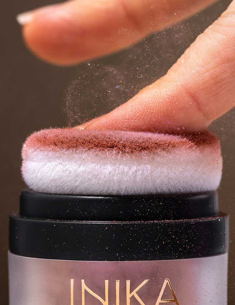 A finger presses onto the soft applicator of INIKA Organic Mineral Blush Puff Pot - Rosy Glow 3g, releasing a fine pink dust.