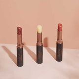 Three INIKA Organic lipsticks, including the INIKA Organic Tinted Lip Balm in Mulberry 3.5g, stand upright in black and gold tubes on a light pink surface against a peach background, shown in mauve, nude, and soft pink shades.