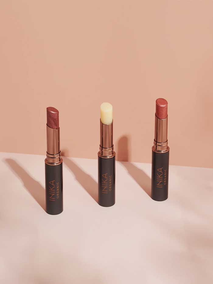 Three INIKA Organic lipsticks, including the INIKA Organic Tinted Lip Balm in Mulberry 3.5g, stand upright in black and gold tubes on a light pink surface against a peach background, shown in mauve, nude, and soft pink shades.