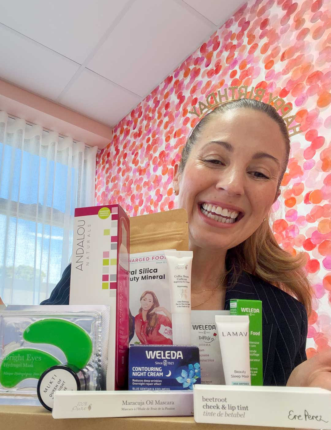 A smiling woman holds Clean Nectarine's My Birthday Box, a skincare gift set featuring creams, mascara, and night cream, in front of a vibrant pink and red polka dot wall.