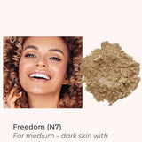 A smiling woman with curly hair and medium-dark skin is shown next to INIKA Organic Loose Mineral Foundation SPF25 8g in shade Freedom (N7), ideal for medium-dark skin with golden or red undertones.