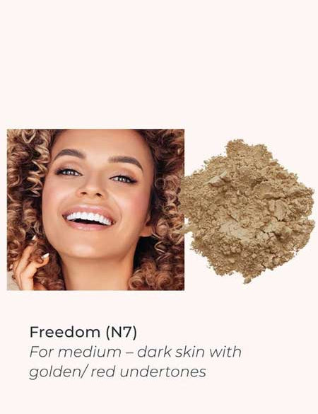 A smiling woman with curly hair and medium-dark skin is shown next to INIKA Organic Loose Mineral Foundation SPF25 8g in shade Freedom (N7), ideal for medium-dark skin with golden or red undertones.