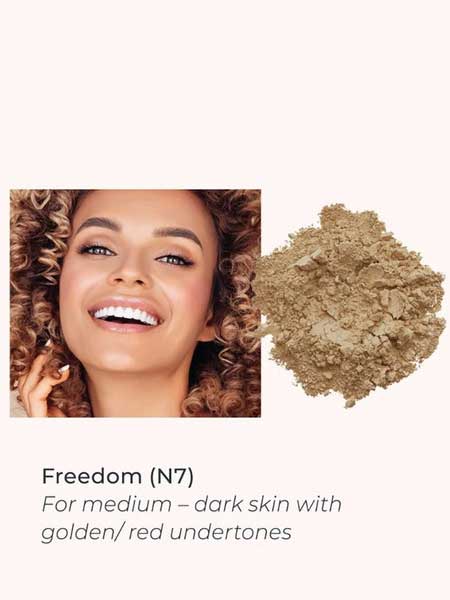 A smiling woman with curly hair and medium-dark skin is shown next to INIKA Organic Loose Mineral Foundation SPF25 8g in shade Freedom (N7), ideal for medium-dark skin with golden or red undertones.