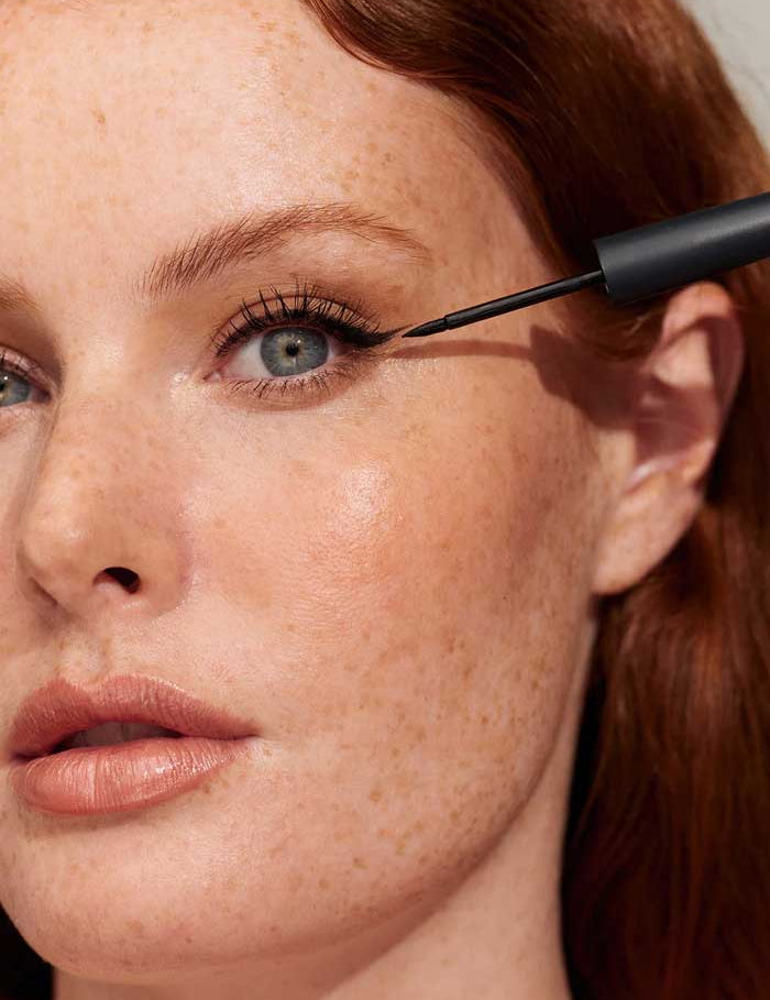 A close-up of a fair-skinned, freckled redhead applying INIKA Organic Liquid Eyeliner 15.9g to her upper eyelid. She wears natural makeup and slightly parts her lips, focusing on a precise, smudge-proof eye look.