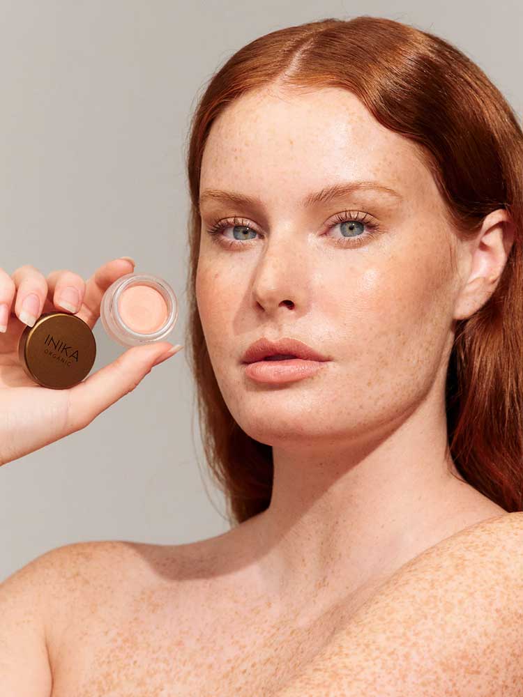 A fair-skinned woman with red hair and freckles holds up a small open pot of INIKA Organic Lip & Cheek Cream Morning 3.5g near her face against a plain background.