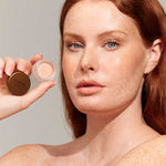 A fair-skinned woman with red hair and freckles holds up a small open pot of INIKA Organic Lip & Cheek Cream Morning 3.5g near her face against a plain background.
