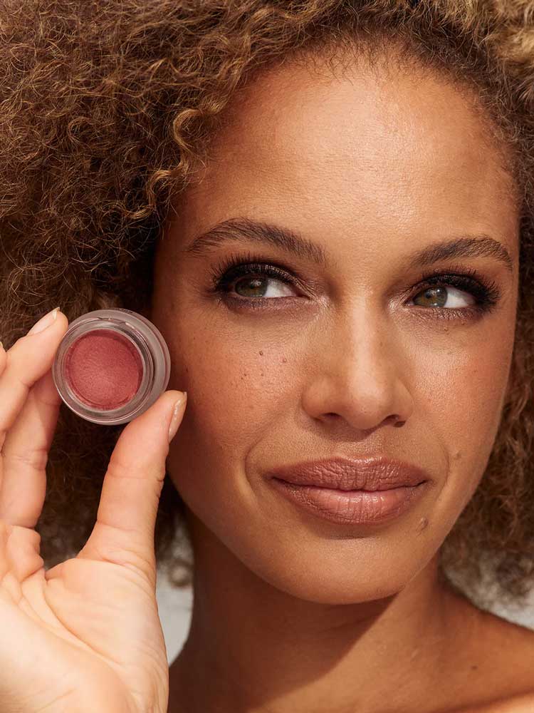 A woman with curly hair holds INIKA Organic Lip & Cheek Cream Petals 3.5g near her face, highlighting her natural makeup look with neutral lipstick and soft eyeshadow.