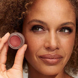 A woman with curly hair holds INIKA Organic Lip & Cheek Cream Petals 3.5g near her face, highlighting her natural makeup look with neutral lipstick and soft eyeshadow.