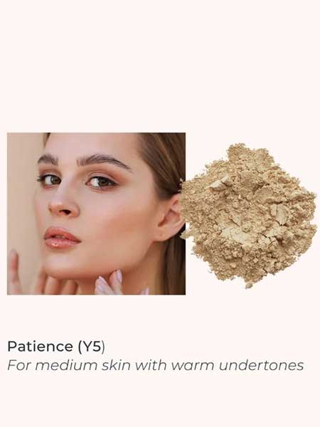 A woman with medium skin touches her face next to a pile of beige INIKA Organic Loose Mineral Foundation SPF25 8g, shade Patience (Y5), for medium skin with warm undertones.
