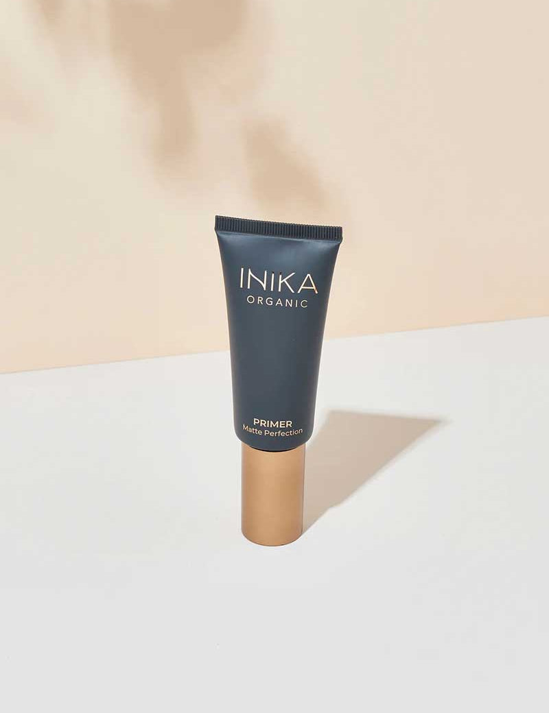A 30ml tube of INIKA Organic Primer Matte Perfection, featuring a dark matte body and gold cap, stands upright on a white surface against a beige background.