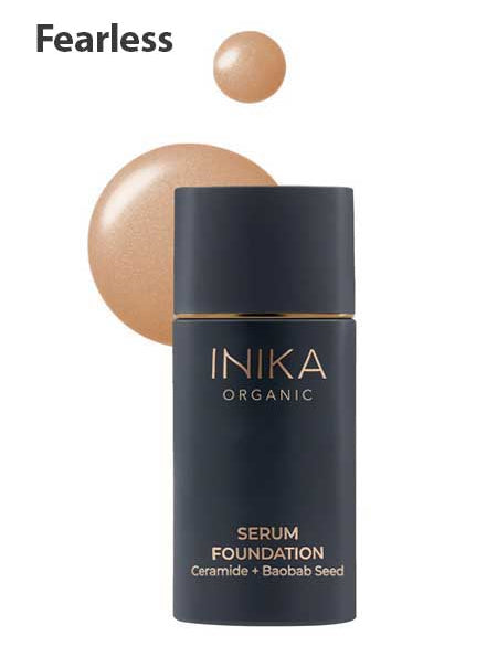A black bottle of INIKA Organic Serum Foundation 25ml, labeled Fearless, with gold text is displayed next to a swatch of the hydrating, anti-ageing makeup's shade, formulated with Ceramide and Baobab Seed.