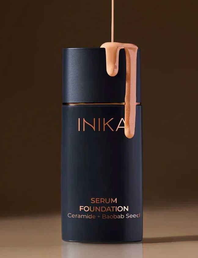 A 25ml bottle of INIKA Organic Serum Foundation with beige liquid drips down its dark navy cap. The label reads "Serum Foundation Ceramide + Baobab Seed." The hydrating foundation is set against a neutral brown background.