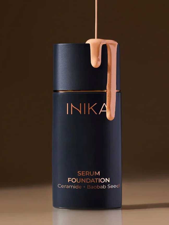 A 25ml bottle of INIKA Organic Serum Foundation with beige liquid drips down its dark navy cap. The label reads "Serum Foundation Ceramide + Baobab Seed." The hydrating foundation is set against a neutral brown background.