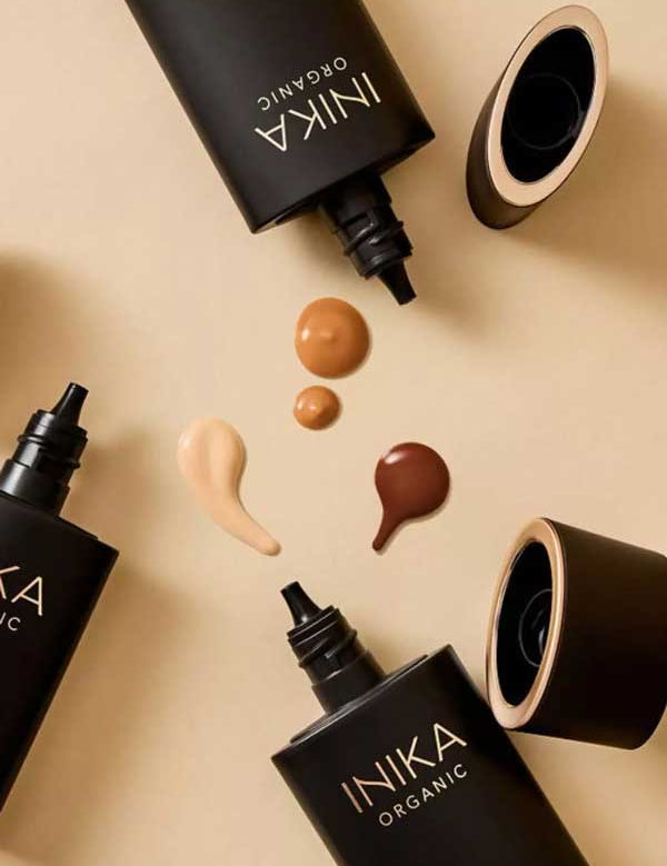 Four black INIKA Organic Serum Foundation 25ml tubes, two uncapped, are arranged on a beige surface with four hydrating foundation swatches in various skin tones displayed at the center.