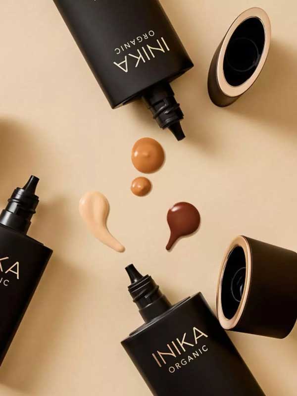 Four black INIKA Organic Serum Foundation 25ml tubes, two uncapped, are arranged on a beige surface with four hydrating foundation swatches in various skin tones displayed at the center.