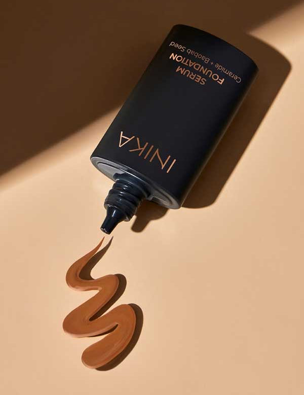 A black tube of INIKA Organic Serum Foundation 25ml with an open nozzle rests on a beige surface, dispensing a swirl of hydrating, rich brown foundation—perfect for sensitive skin.