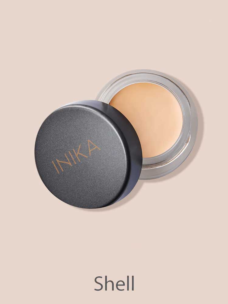 A small round container of INIKA Organic Full Coverage Concealer 3.5g with the lid partially open displays a light beige shade. The word "Shell" is shown below on a matching light beige background.