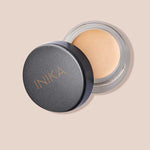 A small round container of INIKA Organic Full Coverage Concealer 3.5g with the lid partially open displays a light beige shade. The word "Shell" is shown below on a matching light beige background.