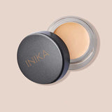A small round container of INIKA Organic Full Coverage Concealer 3.5g with the lid partially open displays a light beige shade. The word "Shell" is shown below on a matching light beige background.
