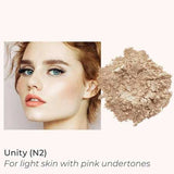 A woman with fair skin and peachy makeup poses beside INIKA Organic Loose Mineral Foundation SPF25 8g in light beige. Text: Unity (N2) for light skin with pink undertones. Non-comedogenic, SPF25 protection.