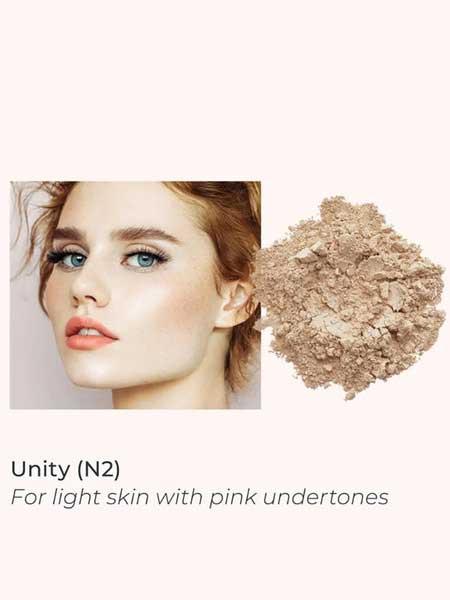 A woman with fair skin and peachy makeup poses beside INIKA Organic Loose Mineral Foundation SPF25 8g in light beige. Text: Unity (N2) for light skin with pink undertones. Non-comedogenic, SPF25 protection.