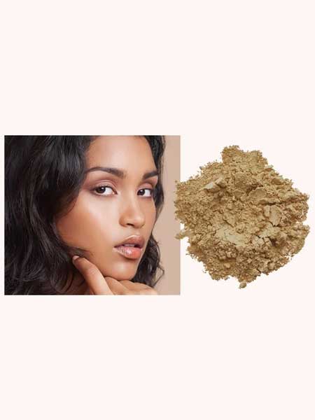 A woman with smooth, glowing skin touches her chin beside a pile of INIKA Organic Loose Mineral Foundation SPF25 8g on a light background.