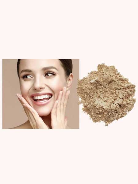 A smiling woman touches her face with both hands beside a pile of INIKA Organic Loose Mineral Foundation SPF25 8g, featuring light beige organic powder on a plain background.