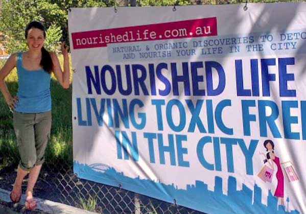 A woman stands next to a large outdoor banner that reads Nourished Life: Living Toxic Free in the City. The banner also displays a website URL and a graphic of a woman holding shopping bags.