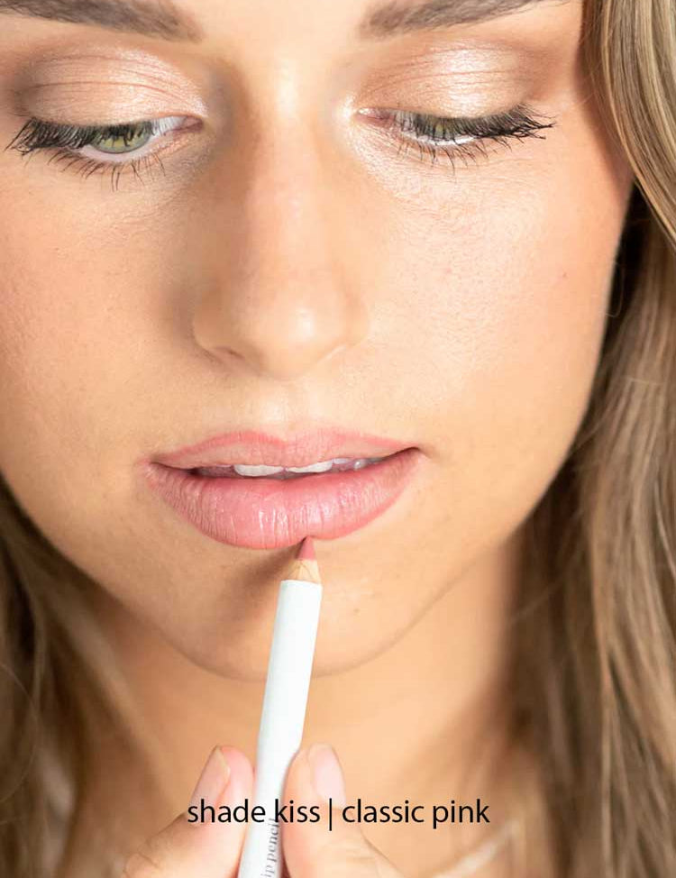A woman with long brown hair and natural makeup applies Ere Perez Acai Lip Pencil 1.1g in the shade Kiss | Classic Pink, a light pink hydrating lip liner, to her lips.