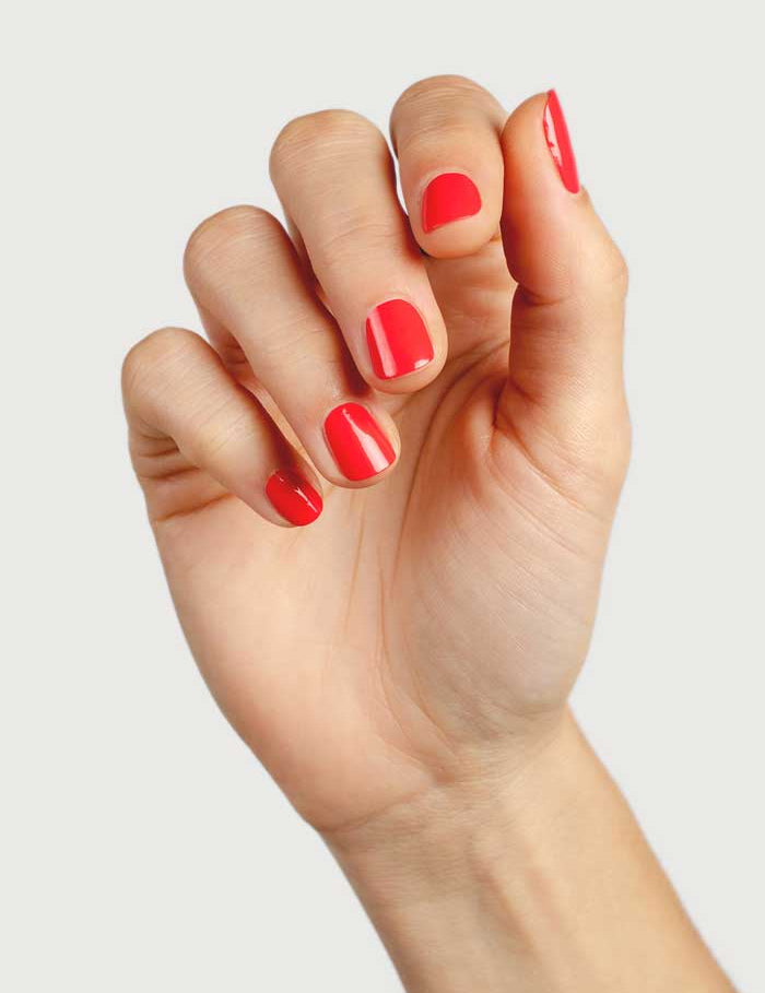 A hand with neatly manicured nails painted in glossy red Sienna Byron Bay Nail Polish - Kiss 10ml is shown against a plain light background. The slightly curled fingers highlight the nails and the natural plant-derived formula.