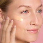 A woman with clear skin applies The Jojoba Company Illuminating Tinted Moisturiser SPF15 50ml to her cheek using her fingers, set against a pink background.