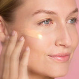 A woman with clear skin applies The Jojoba Company Illuminating Tinted Moisturiser SPF15 50ml to her cheek using her fingers, set against a pink background.