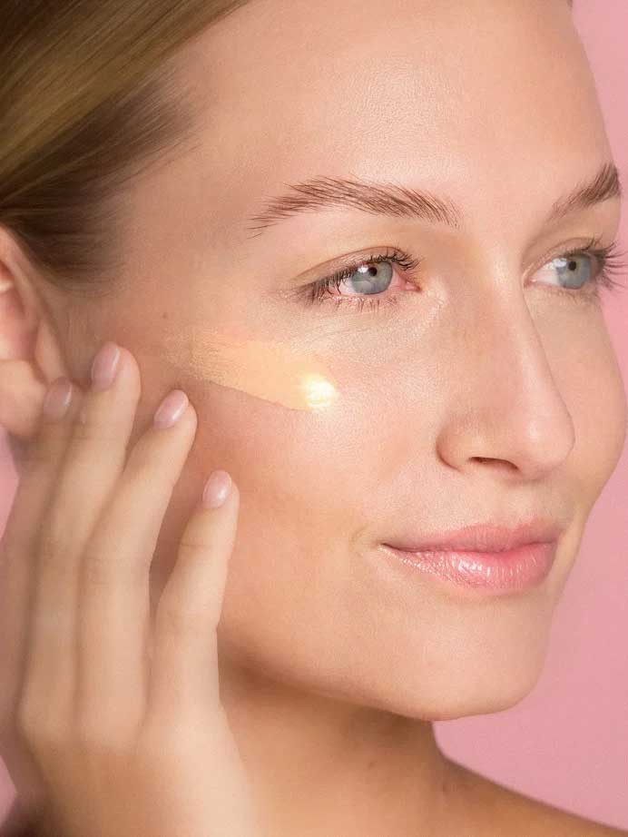 A woman with clear skin applies The Jojoba Company Illuminating Tinted Moisturiser SPF15 50ml to her cheek using her fingers, set against a pink background.