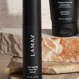 A black bottle of LAMAV Anti-Ageing Face Tan Serum 50ml with hyaluronic acid stands upright on stacked stone slabs, with part of a black Gradual Tanning Moisturiser tube visible in the background.