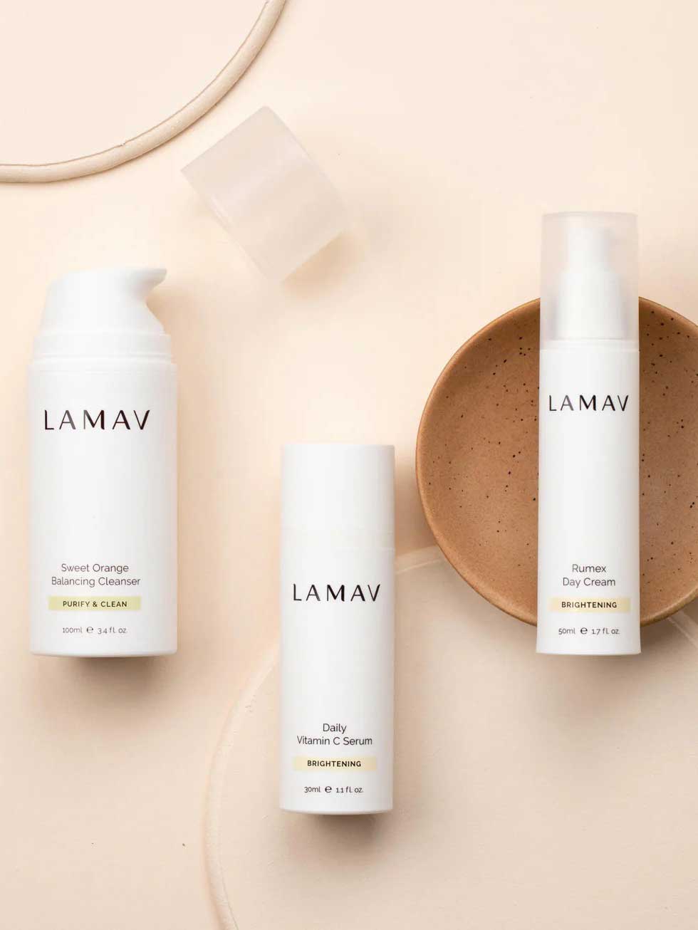 Three white bottles from the LAMAV Brightening Skin Discovery Pack (3 Pack) are displayed on a beige background with a tan ceramic dish.
