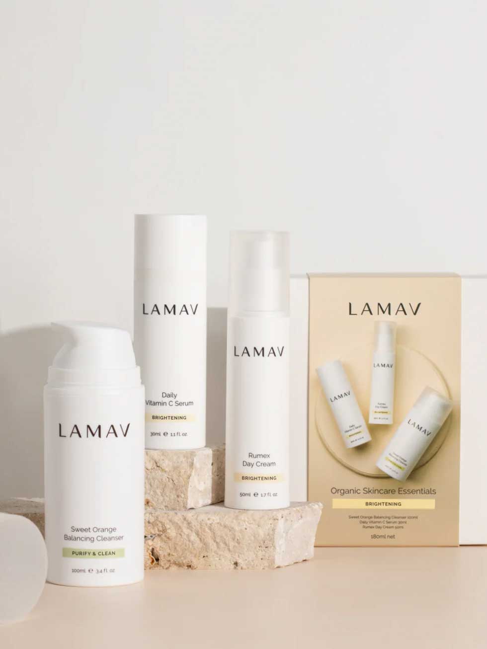 Three LAMAV skincare products from the Brightening Skin Discovery Pack are displayed with minimalist white packaging on a neutral background accented by stone props. A box labeled "Organic Skincare Essentials" features the LAMAV brand.