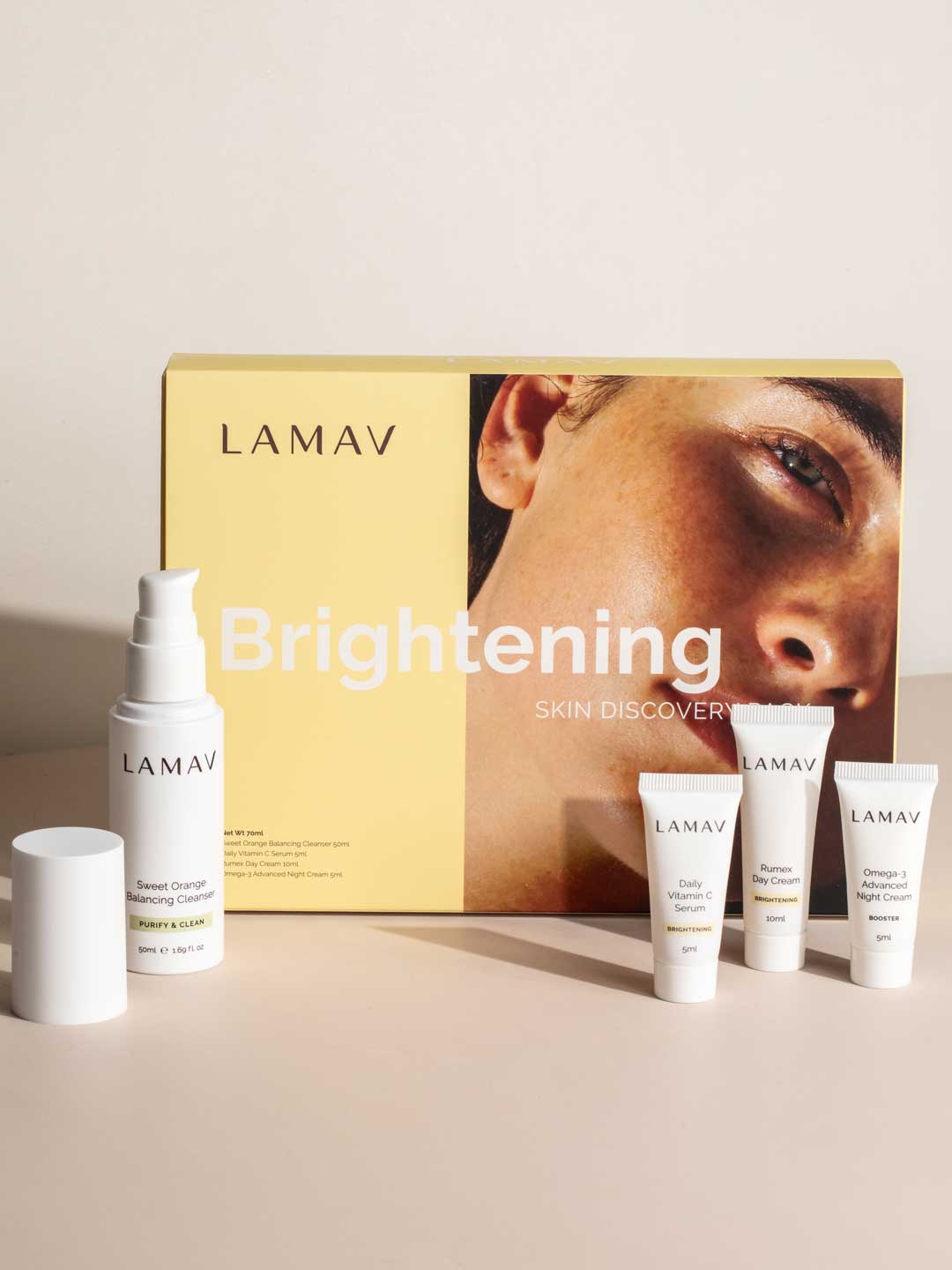 LAMAV Brightening Skin Discovery Pack (4 Pack) by LAMAV shown on a beige surface, with a yellow box featuring a woman's face and four white brightening products—a spray bottle, two small tubes, and a mini bottle. Great for organic skincare lovers.