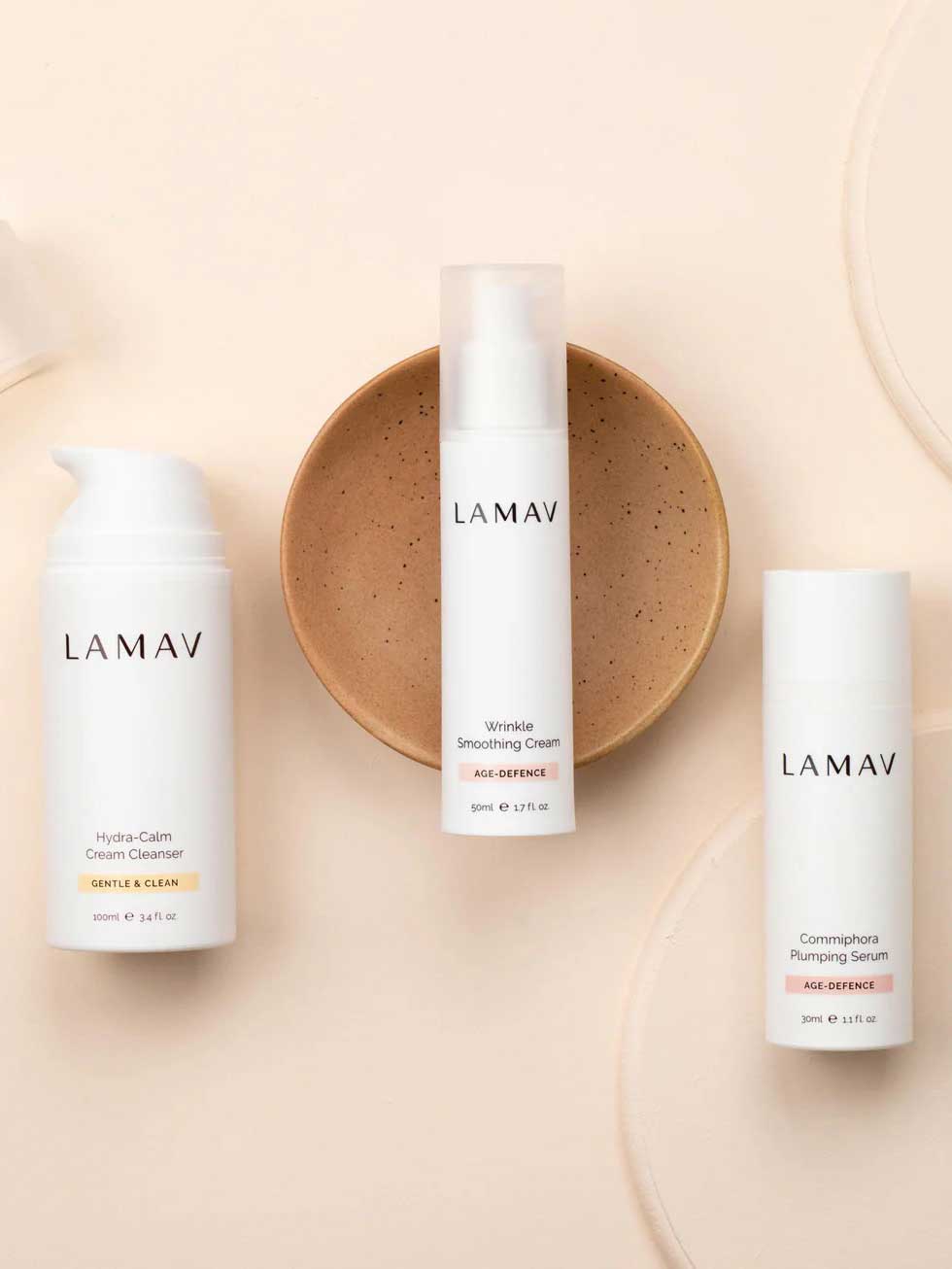 Three LAMAV Age-Defence Skin Discovery Pack bottles are arranged on a beige background—one on a brown dish in the center, with two others beside it. This 3-pack offers an organic vegan skincare routine ideal for anti-ageing.