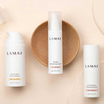 Three LAMAV Age-Defence Skin Discovery Pack bottles are arranged on a beige background—one on a brown dish in the center, with two others beside it. This 3-pack offers an organic vegan skincare routine ideal for anti-ageing.